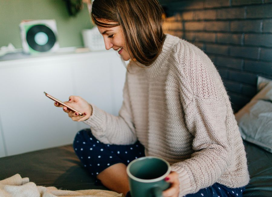 Femme assise sur son lit qui regarde son smartphone en tenant une tasse de café dans l'autre main. 