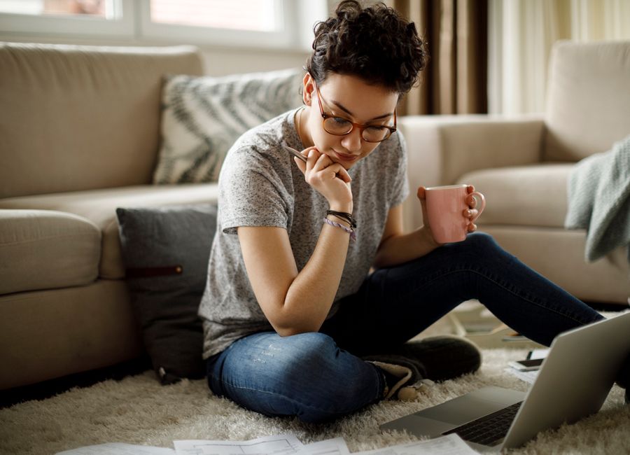 Femme assise par terre face à son ordineur portable, un crayon dans une main et une tasse à café dans l'autre.